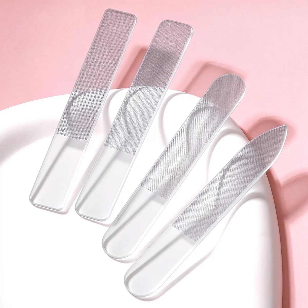 Five transparent glass nail files fanned on a white dish against a pink background