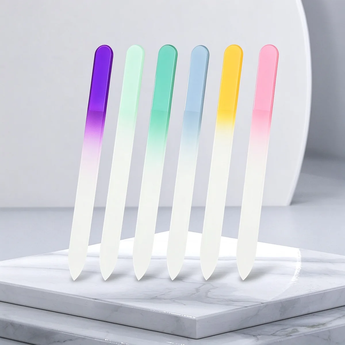 Seven gradient-coloured glass nail files standing on a white marble slab against a pink background