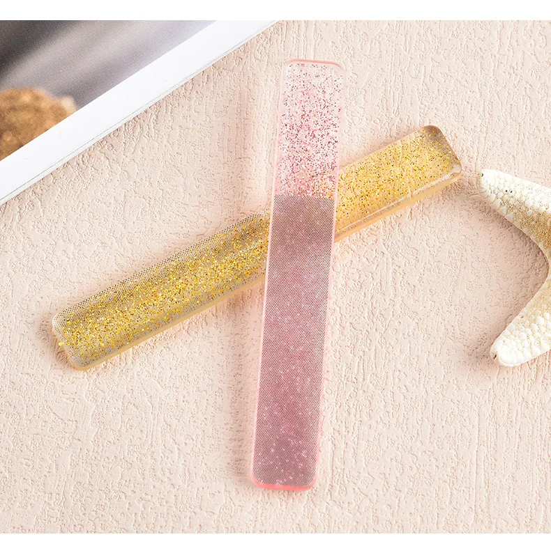 Two glitter glass nail files, one gold and one pink, crossed on a beige linen cloth with shells